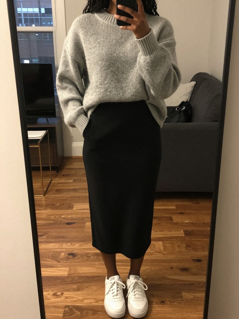 iPhone mirror selfie of a black woman in an oversized grey sweater over a black midi skirt with white sneakers, hands in sweater pockets, cropped at NECK so face doesn't show, in a city apartment, warm moody lighting, iPhone photo quality.