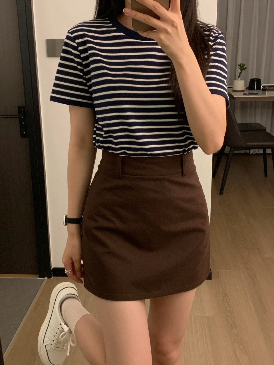 iPhone mirror selfie of a woman in a navy striped tee tucked into a chocolate brown mini skirt with white sneakers, adjusting cap brim, cropped at NECK so face doesn't show, in a city apartment, warm moody lighting, iPhone photo quality.