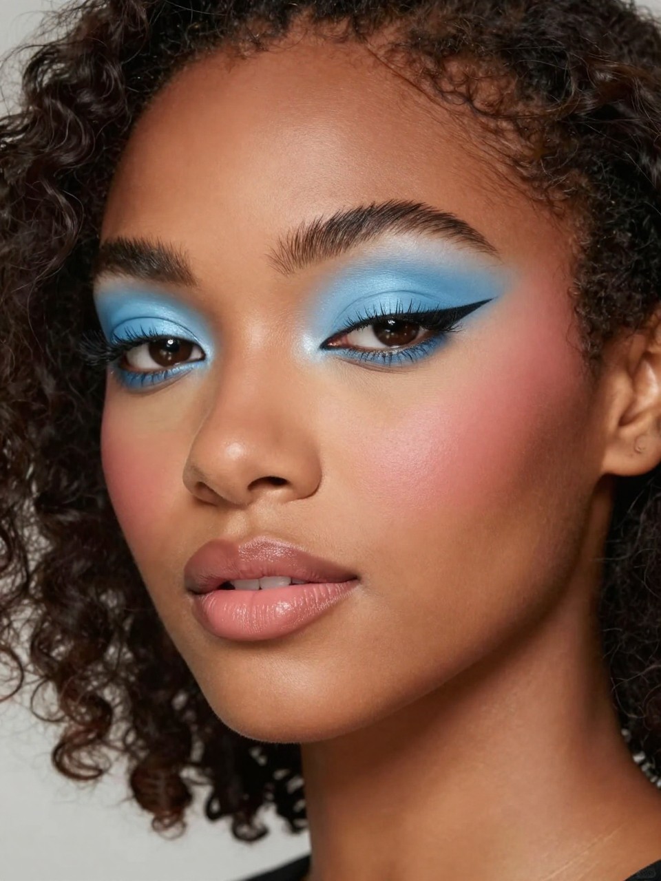 IMAGE_PROMPT: Super realistic close-up beauty portrait of a Black woman wearing baby blue matte eyeshadow across the lids with a short soft black winged liner, volumized lashes, smooth balanced skin, soft pink blush, peachy nude lipstick, and softly arched brows, shot from a three-quarter face angle, with diffused studio lighting that clearly reveals the liner shape and blue shadow color, realistic skin texture, shoulder-length soft curls, minimal blurred background, feminine modern beauty photography, latest iPhone photo quality (no watermarks on images).