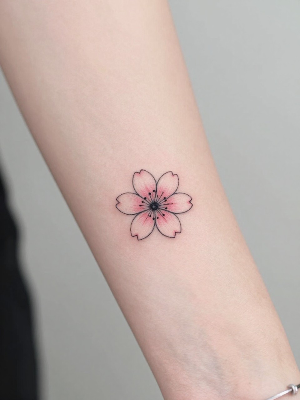 design-focused close-up of a small Japanese sakura tattoo on the wrist, tiny blossom with delicate petals, clean black ink with soft blush pink shading, fine line detail, realistic skin texture, bracelet nearby, minimal blurred background, modern tattoo inspiration photo (no watermarks on images)