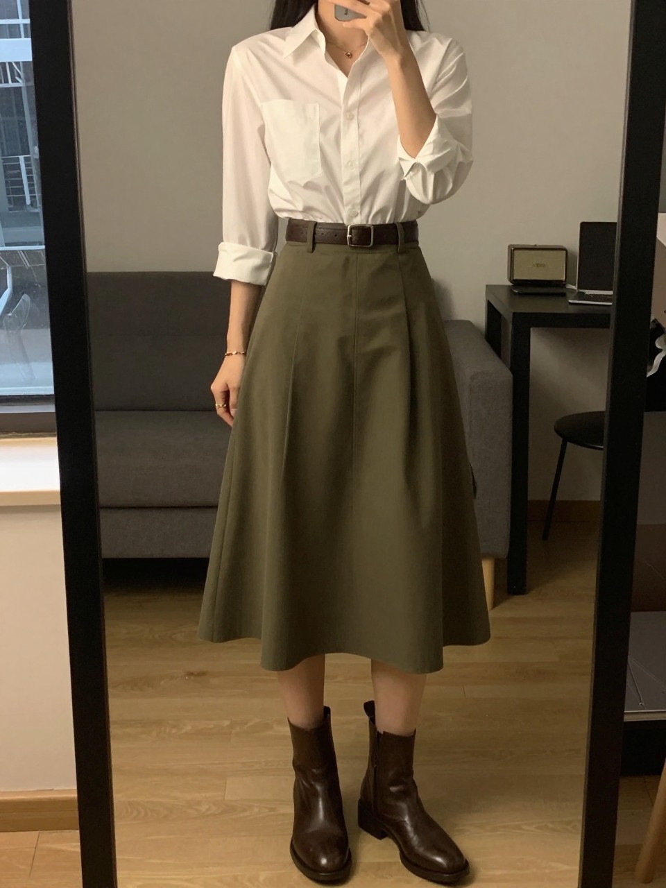 iPhone mirror selfie of a woman in a white button-down tucked into an olive green A-line skirt with a brown belt and brown ankle boots, one hand on belt, cropped at NECK so face doesn't show, in a city apartment, warm moody lighting, iPhone photo quality.