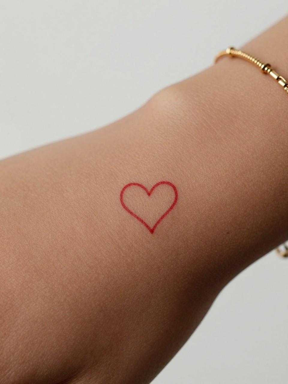 extreme close-up of a tiny red heart outline tattoo on deep brown skin at the inner wrist, clean red ink, crisp fine line, very small minimalist design, realistic skin texture, neat neutral manicure, delicate gold bracelet, design-focused close-up, soft minimal background, modern feminine tattoo inspiration photography (no watermarks on images)"