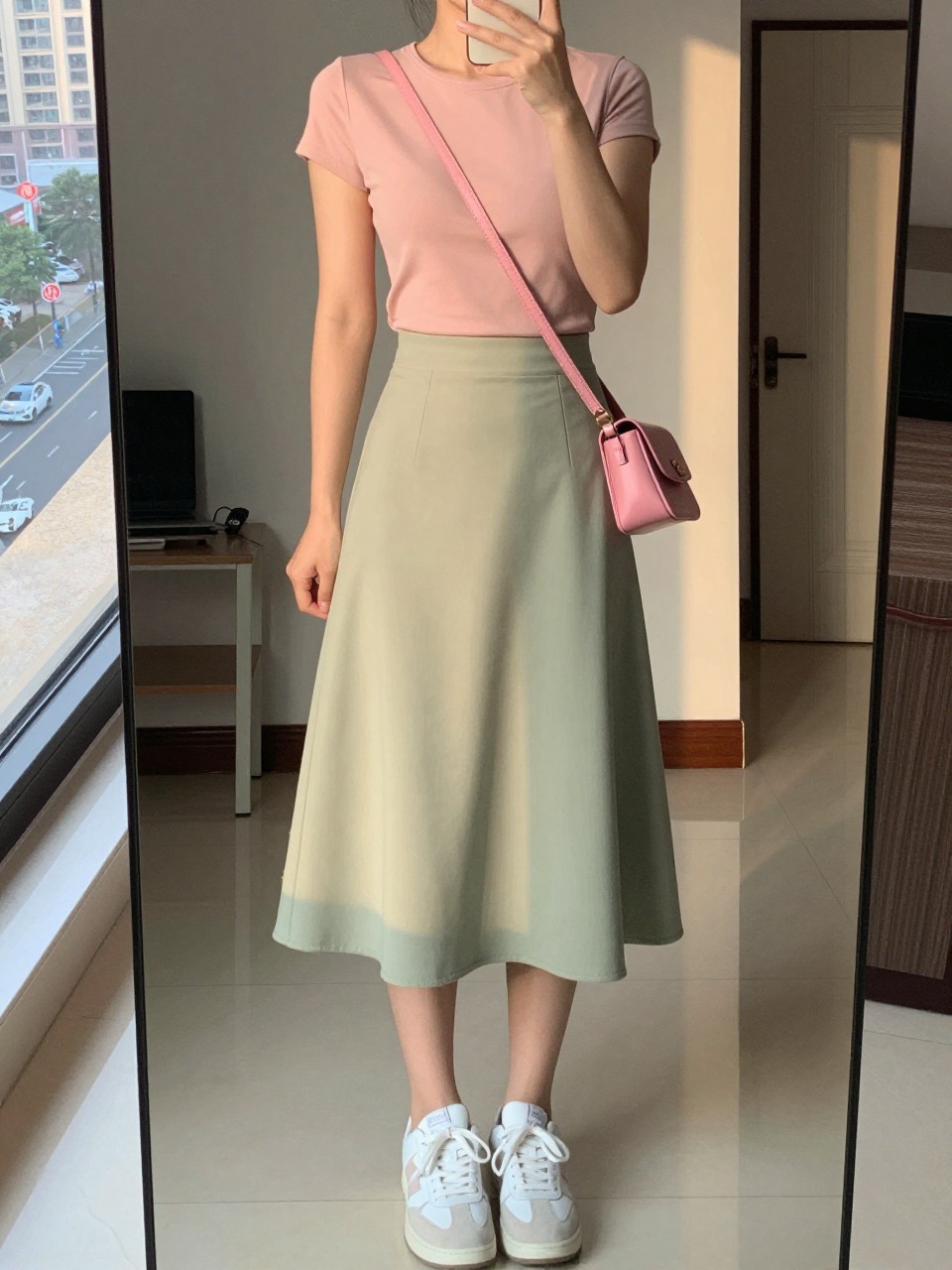 iPhone mirror selfie of a woman in a blush pink top tucked into a sage green A-line skirt with white sneakers, holding a pink crossbody bag, cropped at NECK so face doesn't show, in a city apartment, evening golden hour, iPhone photo quality.