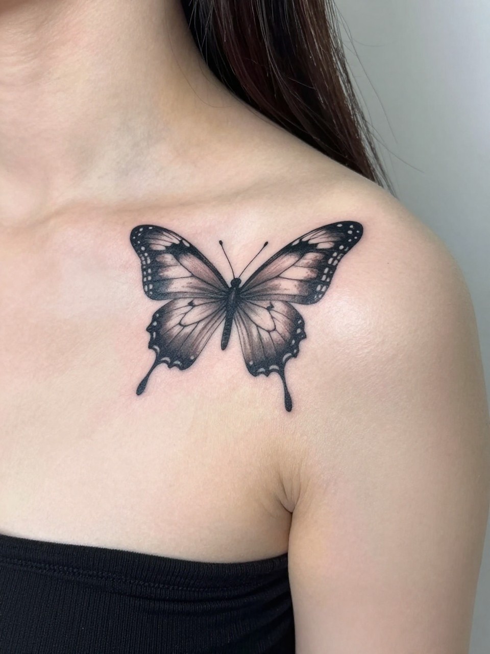 "Angled shot from above: a butterfly tattoo flying along the line of the right collarbone, its wings breaking the plane of the shoulder and chest. Asymmetric and dynamic."