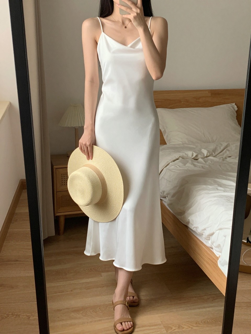 iPhone mirror selfie of a woman in a white slip dress and tan sandals, holding a wide-brim hat, cropped at the neck so face doesn't show, in a feminine-themed bedroom, warm moody lighting, iPhone photo quality.