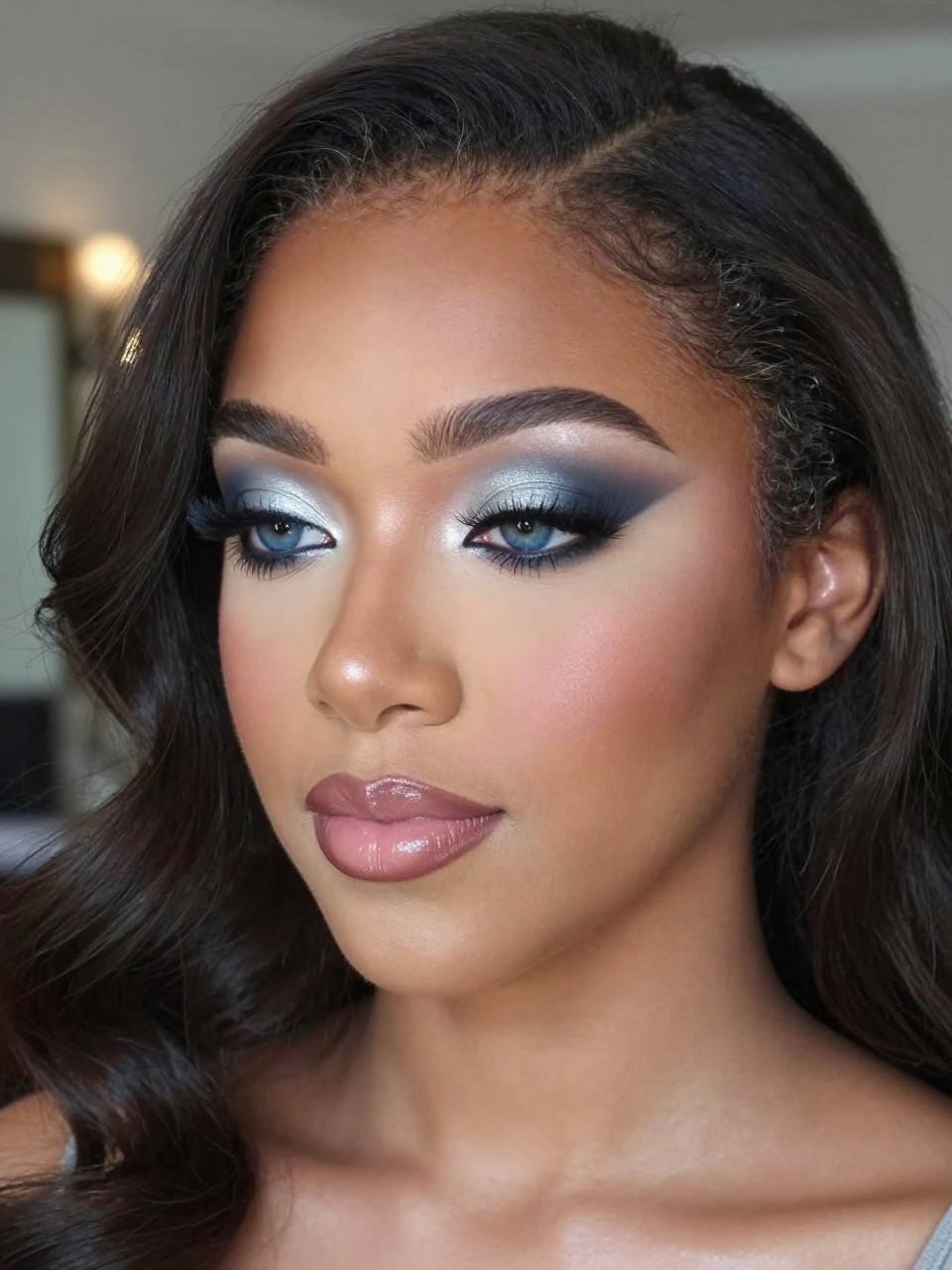 Super realistic close-up beauty portrait of a black teenage woman with blue eyes wearing lovely prom makeup, featuring gray-blue eyeshadow across the lids, silver-taupe crease blending, soft charcoal outer-corner shading, defined volumized lashes, cool pearl highlight, soft nude-pink blush, nude mauve glossy lips, refined brows, realistic skin texture with a silky satin glow, shot from a three-quarter portrait angle, with cool elegant lighting that clearly reveals the gray-blue tones, blue eyes, and mauve gloss, softly styled waves, blurred evening formal background, feminine modern beauty photography, latest iPhone photo quality.