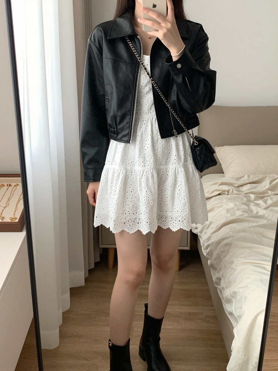 mirror selfie of a woman wearing the exact outfit described above, clearly showing a cropped black leather jacket with a neat fitted shape, white eyelet mini dress with soft texture, black ankle boots, small black quilted crossbody bag, and a delicate silver bracelet, holding a phone in front of a mirror, cropped at NECK so the face does not show, with a relaxed casual selfie pose, in a cozy bedroom mirror setup, natural window light, including soft pillows on an unmade bed and a jewelry tray on the vanity, latest iPhone photo quality.