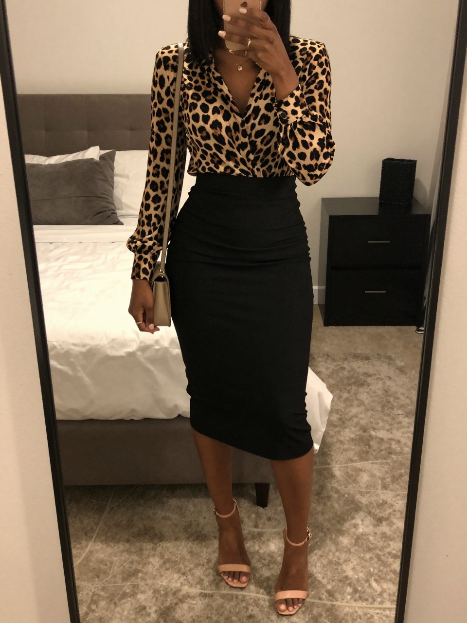 iPhone mirror selfie of a black woman in a leopard print blouse tucked into a black midi skirt with nude heeled sandals, holding a small shoulder bag, cropped at NECK so face doesn't show, in a feminine-themed bedroom, warm moody lighting, iPhone photo quality.