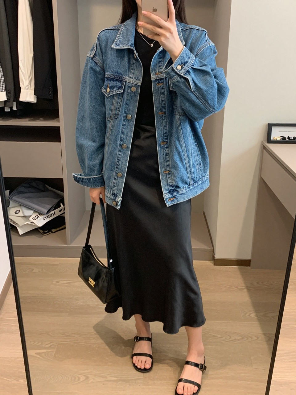 mirror selfie of a woman wearing the exact outfit described above, clearly showing a medium blue oversized denim jean jacket, soft black fitted tee, black satin midi slip skirt, black leather strappy flat sandals, and a small smooth black shoulder bag, holding a phone in front of a mirror, cropped at NECK so the face does not show, with a relaxed casual selfie pose, in a walk-in closet mirror, soft indoor lighting, including an open closet with clothes visible and a jewelry tray on the vanity, latest iPhone photo quality.