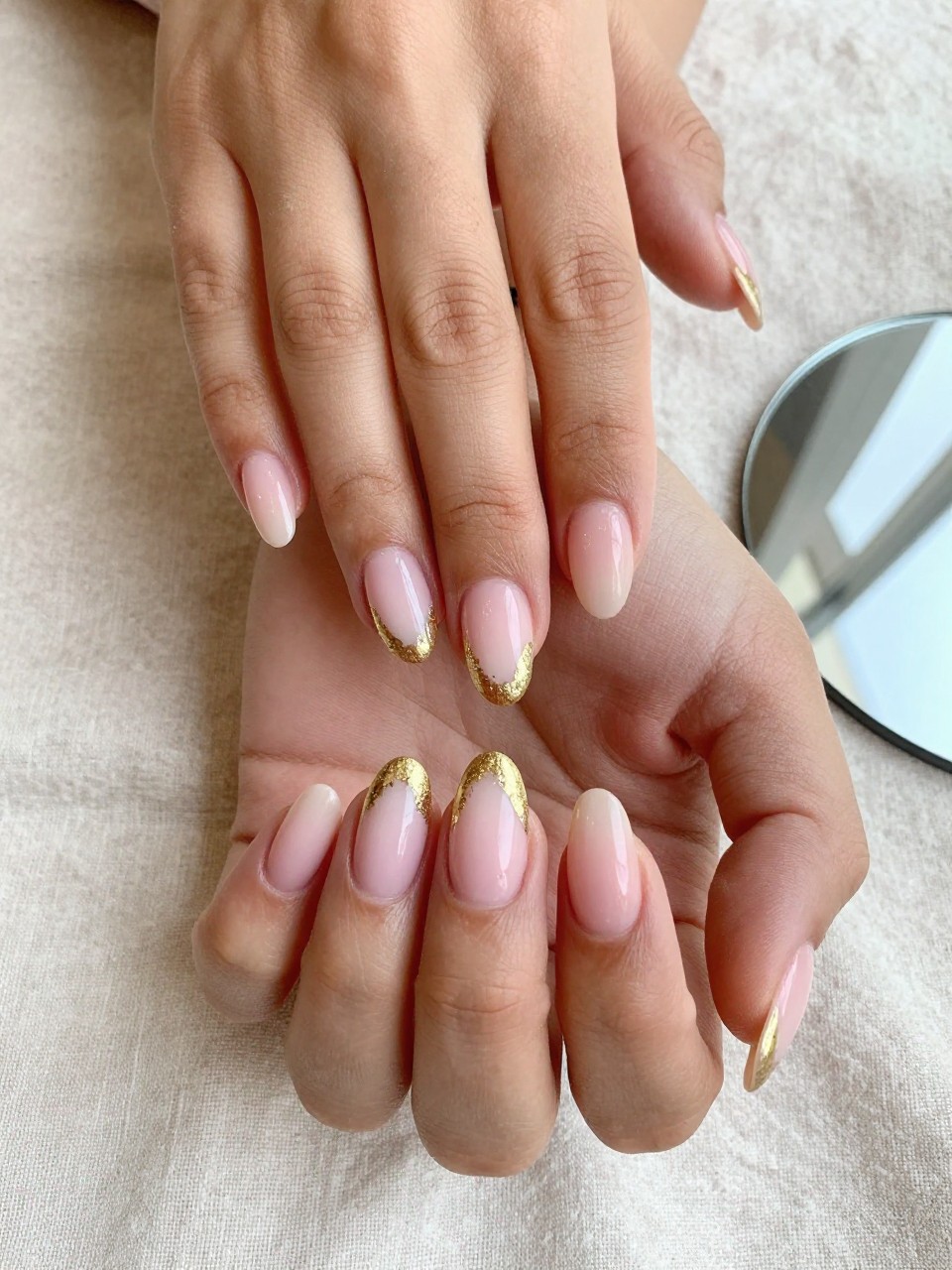 90-degree overhead top-down view of two feminine black hands with almond medium nails.
The manicure features a soft pink ombré base with gold foil pressed along the outer edges of the nails.
Include reflective metallic detail with a glossy gel finish.

Hands are posed fingers gently spread.

Scene set on a soft linen cloth with a small mirror nearby.

Bright natural daylight with soft shadows, hyper-realistic lifestyle photography, sharp focus on nails, natural skin texture, realistic hands, no extra fingers.