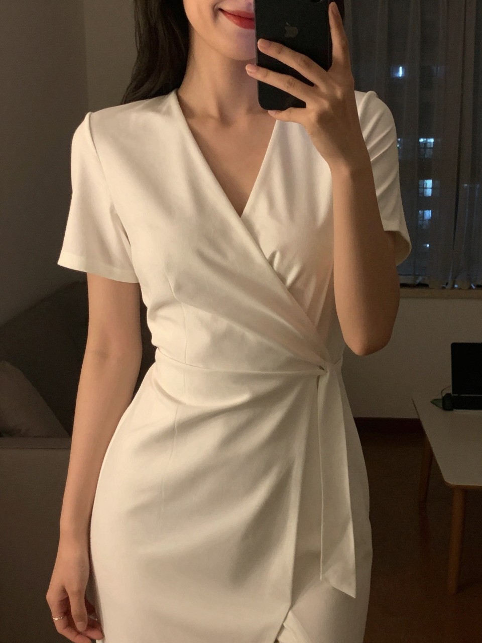 iPhone mirror selfie of a woman in a white wrap dress with a red lip, smiling, cropped at the neck so face doesn't show, in a city apartment, warm moody lighting, iPhone photo quality.