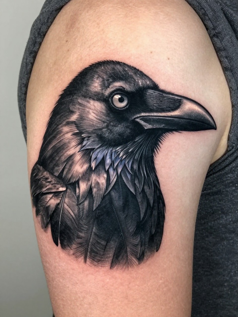  "macro shot of a dark black crow head portrait tattoo on the upper arm, sharp beak, focused eye, detailed layered feathers, black and grey shading, bold realistic blackwork, tight crop, realistic skin texture, soft knit top edge visible, minimal neutral background, modern tattoo inspiration image (no watermarks on images)"