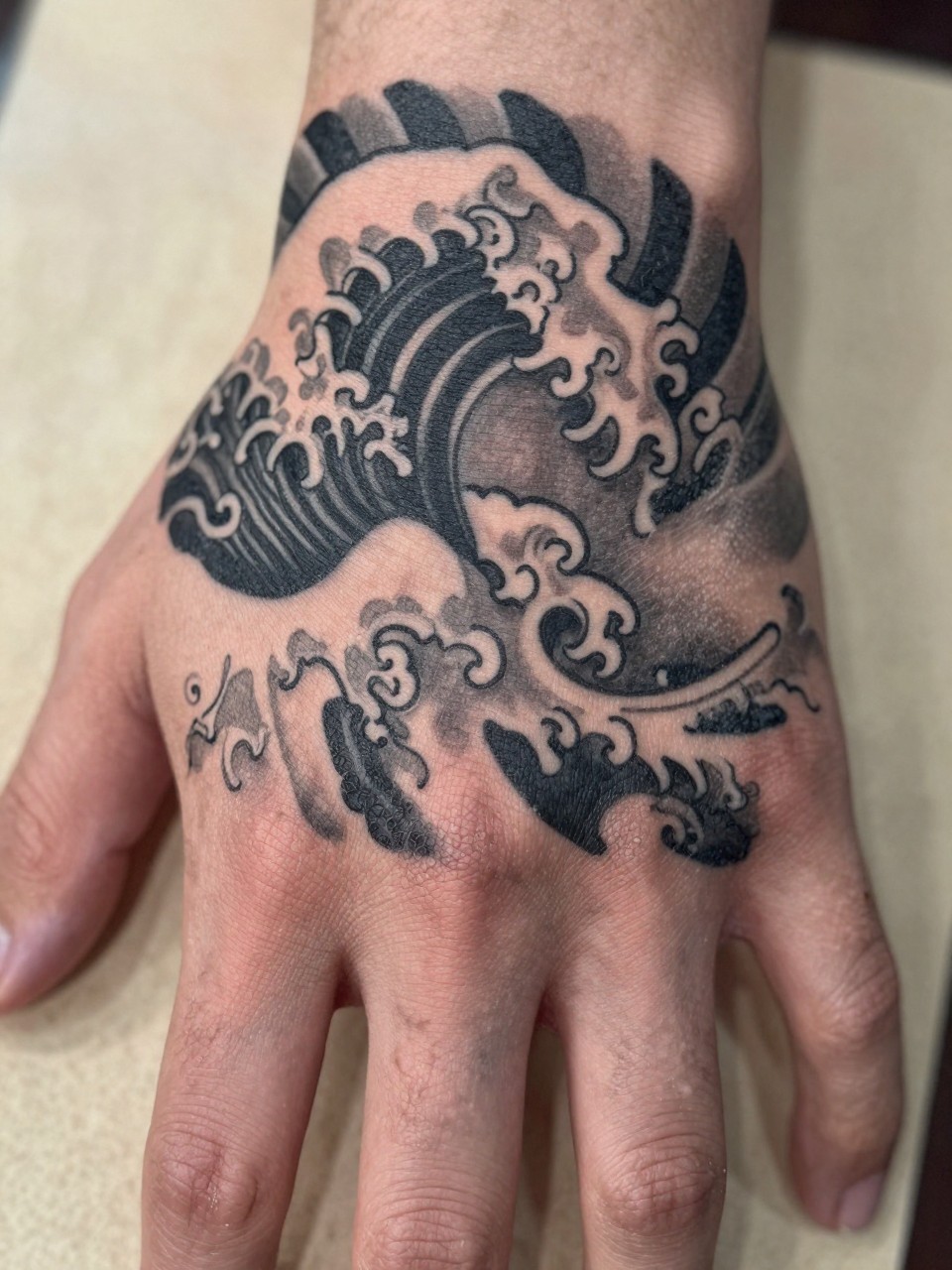 macro shot of a traditional Japanese wave tattoo across the back of the hand, curling wave forms, black and grey ink, bold traditional linework, soft shading, realistic skin texture, design-first tattoo image, soft neutral background, tattoo inspiration image (no watermarks on images)