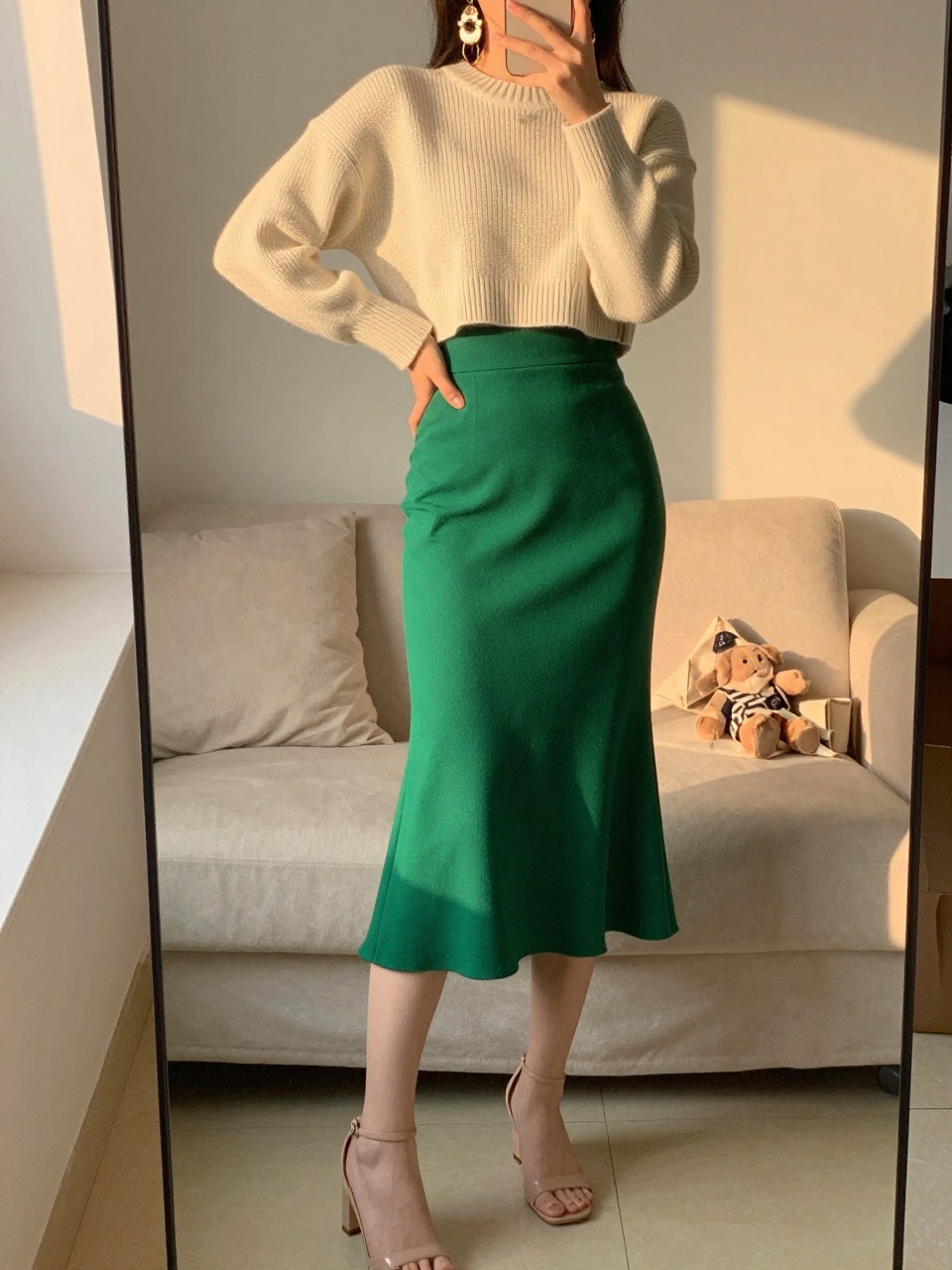 iPhone mirror selfie of a woman in a cream cropped sweater and high-waisted emerald green midi skirt with nude heeled sandals and statement earrings, standing with one hand on hip, cropped at NECK so face doesn't show, in a feminine-themed bedroom, evening golden hour, iPhone photo quality.