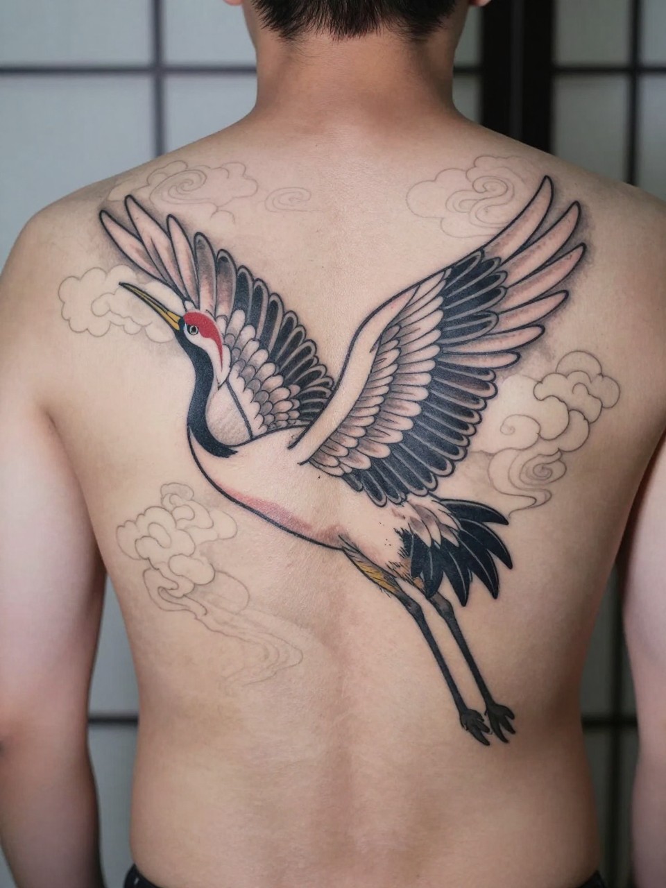 design-focused close-up of a full back Japanese crane tattoo on a man, elegant crane with spread wings, cloud details across the back, black and grey ink, clean traditional linework, soft shading, realistic skin texture, minimal blurred background (no watermarks on images)