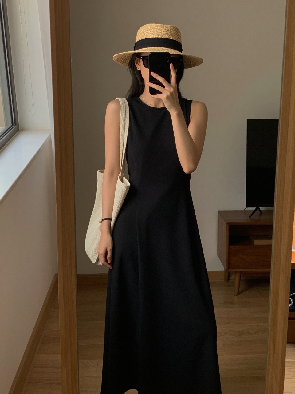 iPhone mirror selfie of a woman in a sleeveless maxi with a straw hat and tote, putting on sunglasses, cropped at NECK so face doesn't show, in a city apartment, warm moody lighting, iPhone photo quality.