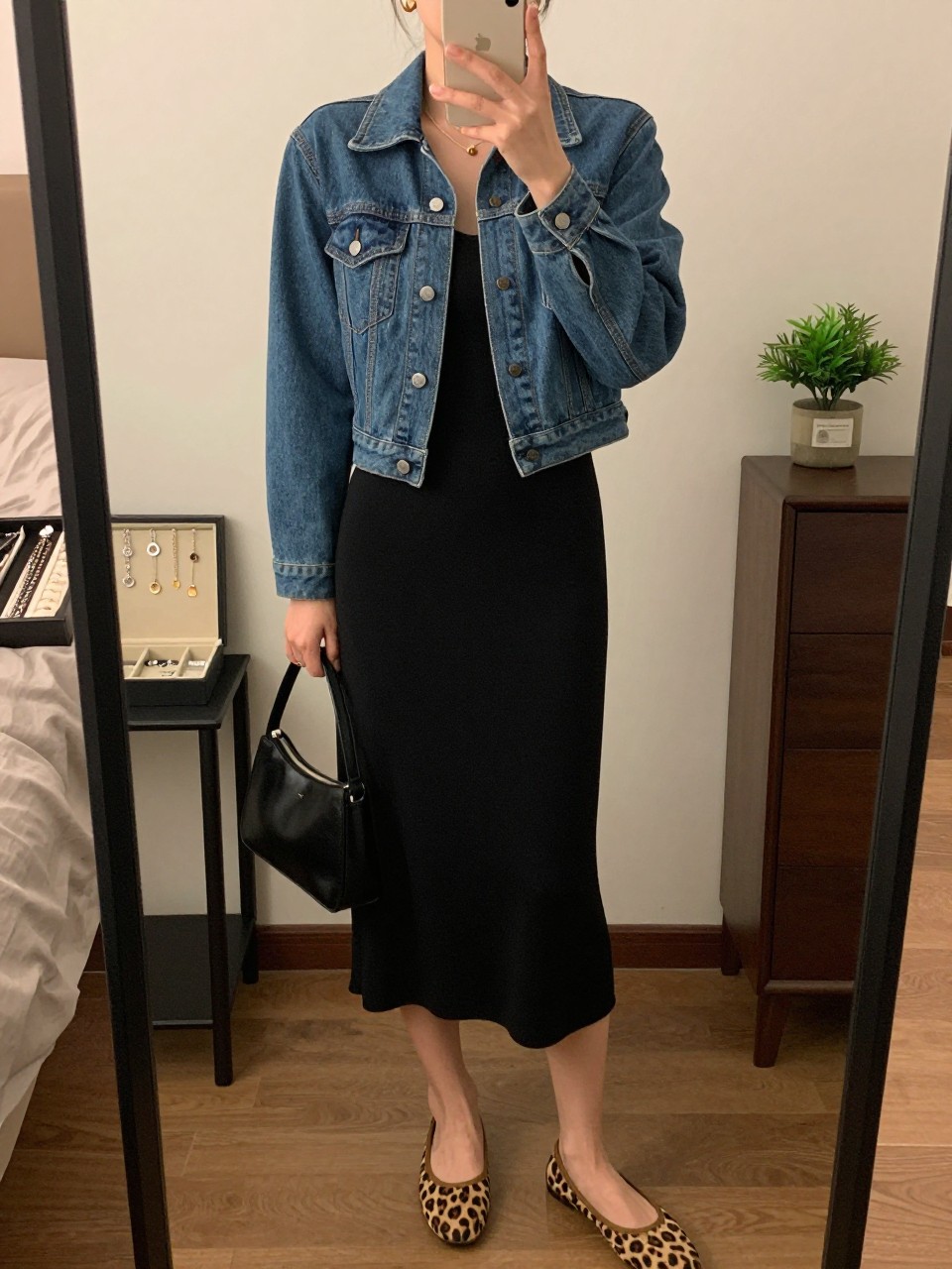 mirror selfie of a woman wearing the exact outfit described above, clearly showing a fitted blue denim jean jacket with a waist-skimming cut, black knit midi dress in soft stretch fabric, leopard print ballet flats, black mini handbag, and small gold hoop earrings, holding a phone in front of a mirror, cropped at NECK so the face does not show, with a relaxed casual selfie pose, in a cozy bedroom mirror setup, soft indoor lighting, including a jewelry tray on the vanity and a small plant on a dresser, latest iPhone photo quality.