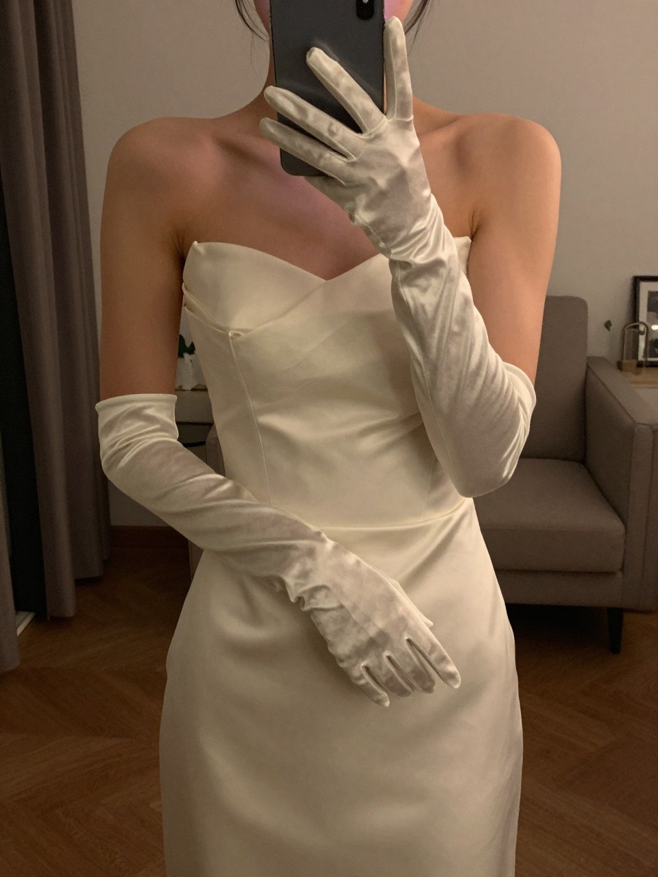 iPhone mirror selfie of a woman in a gown with satin gloves, adjusting her glove, cropped at NECK so face doesn't show, in a city apartment, warm moody lighting, iPhone photo quality.