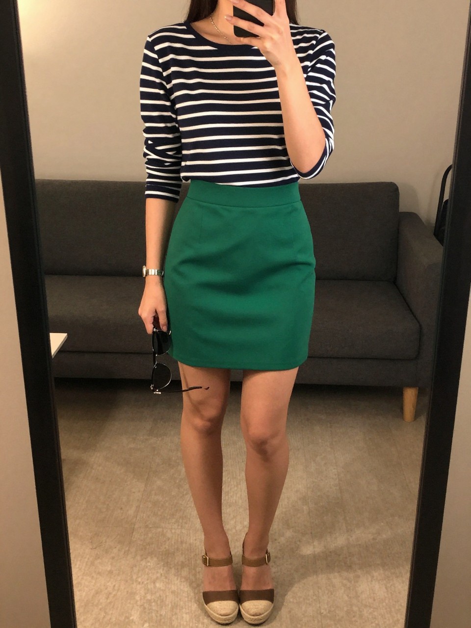iPhone mirror selfie of a woman in a navy striped top tucked into a kelly green mini skirt with espadrille wedges, holding sunglasses, cropped at NECK so face doesn't show, in a city apartment, warm moody lighting, iPhone photo quality.