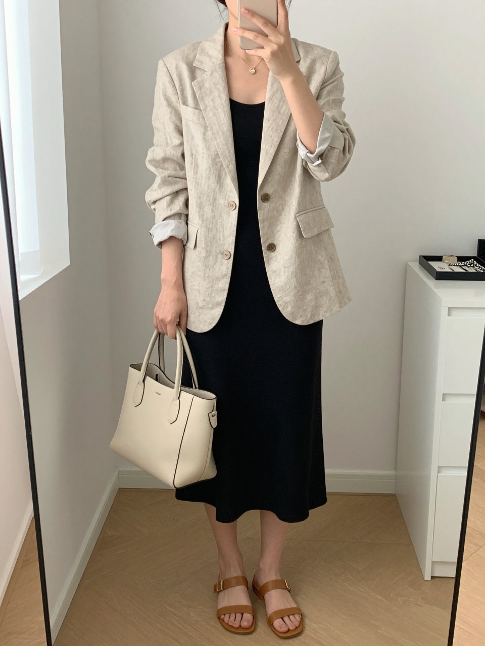 mirror selfie of a woman wearing the exact outfit described above, clearly showing an oatmeal linen blazer, a black soft midi dress, tan low-heel leather sandals, a structured cream leather handbag, and a glossy neutral hair clip, holding a phone in front of a mirror, cropped at NECK so the face does not show, with a relaxed casual selfie pose, in a minimal dressing room, natural window light, including handbag hanging on a door handle and jewelry tray on the vanity, latest iPhone photo quality. (no watermarks on images)