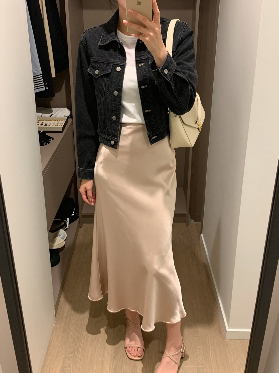 mirror selfie of a woman wearing the exact outfit described above, clearly showing a fitted black denim jean jacket, white fitted crew-neck tee, beige satin midi slip skirt, nude strappy sandals, and a small cream shoulder bag, holding a phone in front of a mirror, cropped at NECK so the face does not show, with a relaxed casual selfie pose, in a walk-in closet mirror, soft indoor lighting, including an open closet with clothes visible and a jewelry tray on the vanity, latest iPhone photo quality.
