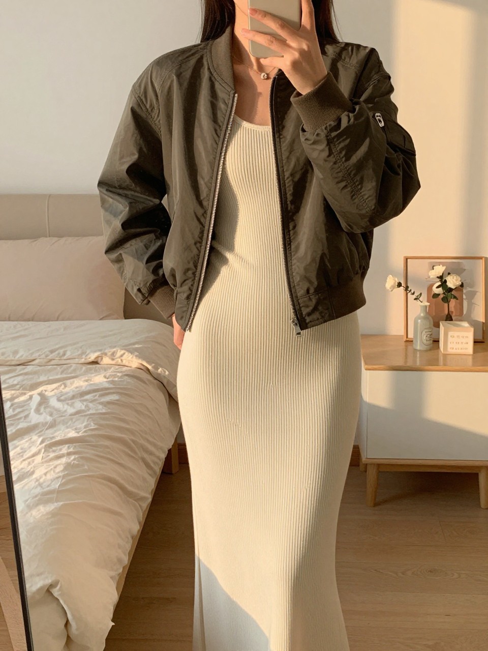 iPhone mirror selfie of a woman in a ribbed maxi with a bomber jacket, hand in pocket, cropped at NECK so face doesn't show, in a feminine-themed bedroom, evening golden hour, iPhone photo quality.