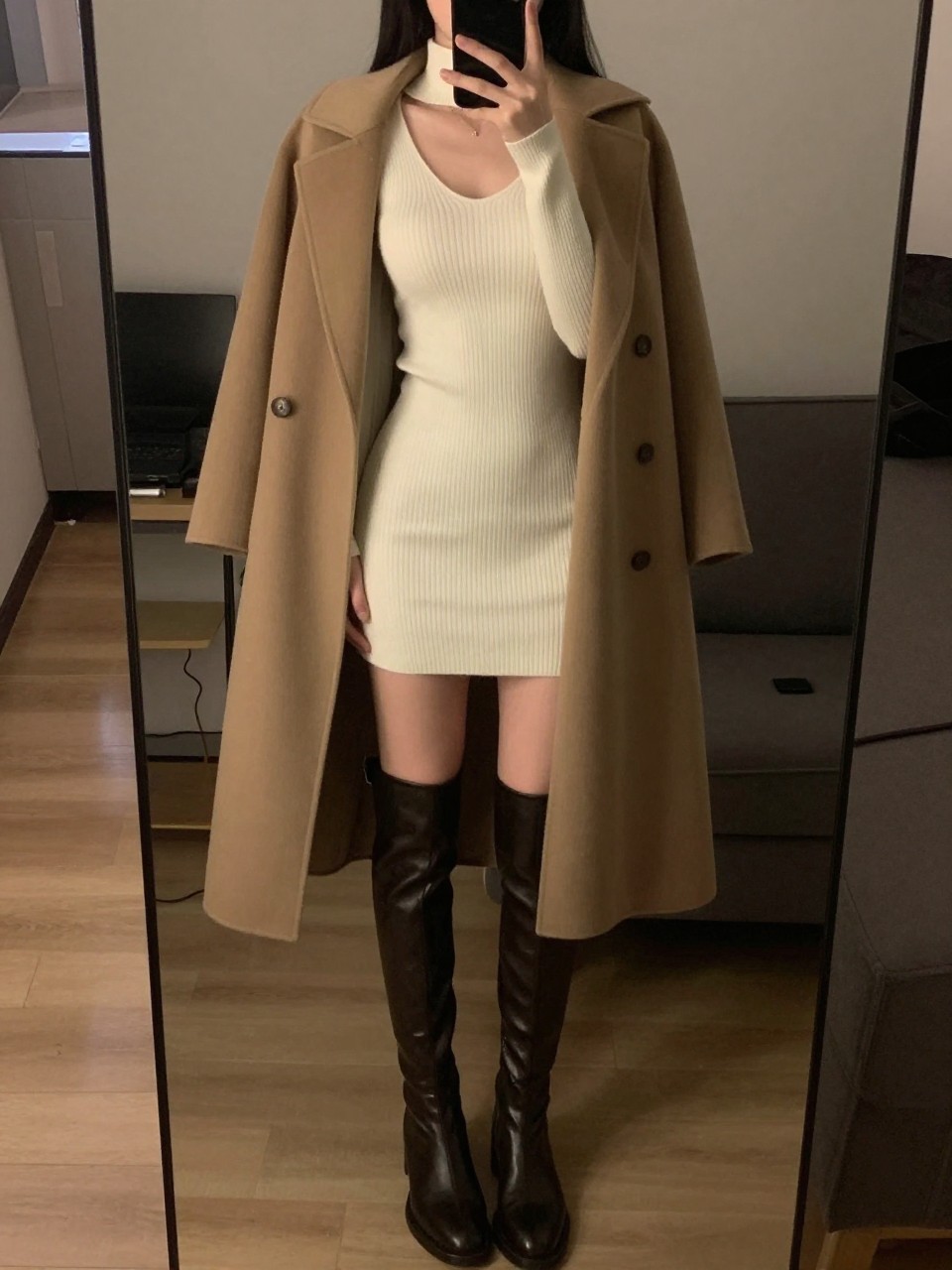 iPhone mirror selfie of a woman in a white knit dress and knee-high boots, wrapped in a coat, cropped at the neck so face doesn't show, in a city apartment, warm moody lighting, iPhone photo quality.