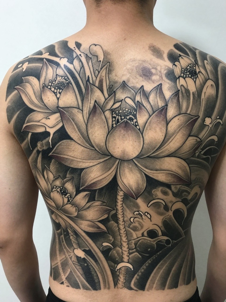 "close-up of a full back Japanese lotus and water tattoo on a man, large lotus blooms with flowing water accents across the back, black and grey ink, fine traditional linework, soft shading, realistic skin texture, design-focused tattoo visibility, minimal background (no watermarks on images)"