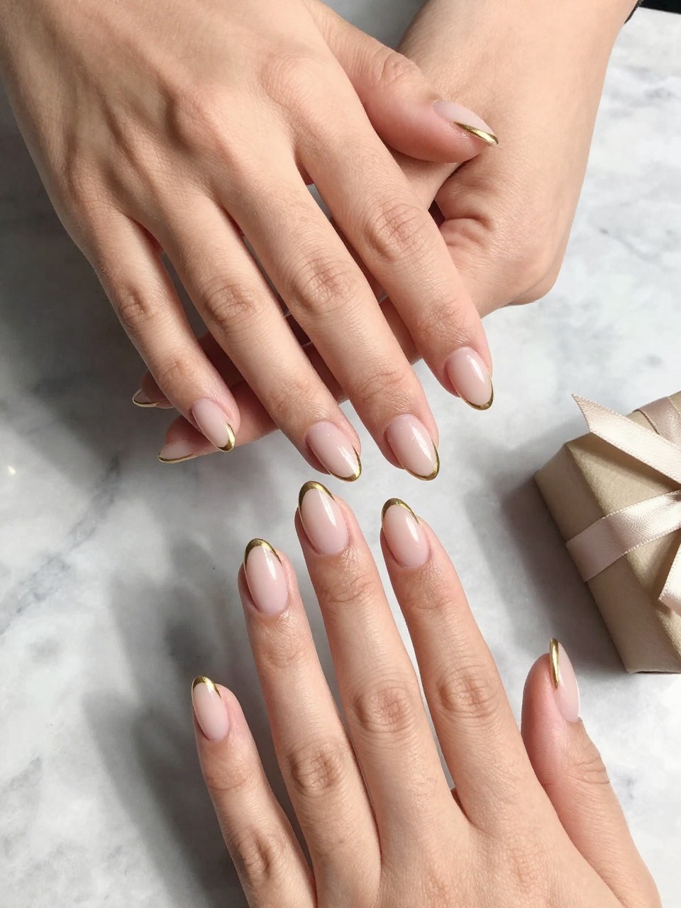 90-degree overhead top-down view of two feminine hands with almond medium nails.
The manicure features a soft nude base with thin gold outline French tips along the edges.
Include subtle metallic shine with a glossy gel finish.

Hands are posed fingers gently spread.

Scene set on a marble surface with a small wrapped gift nearby.

Bright natural daylight with soft shadows, hyper-realistic lifestyle photography, sharp focus on nails, natural skin texture, realistic hands, no extra fingers.