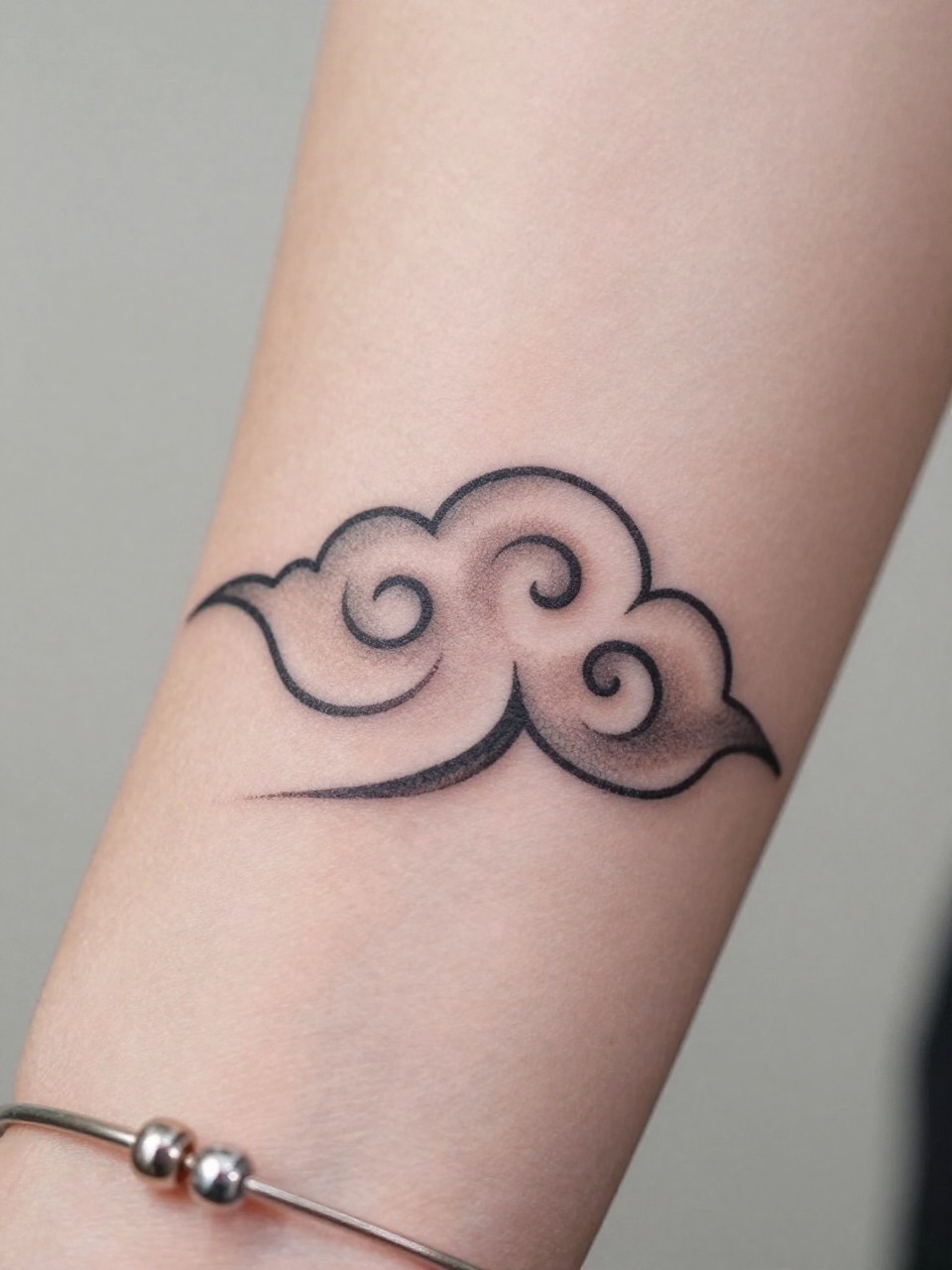 macro shot of a small Japanese cloud tattoo on the wrist, clean black and grey ink, traditional cloud curves, fine crisp linework, soft shading, realistic skin texture, delicate bracelet nearby, tight crop, design-first tattoo composition, soft neutral background, tattoo inspiration image (no watermarks on images)