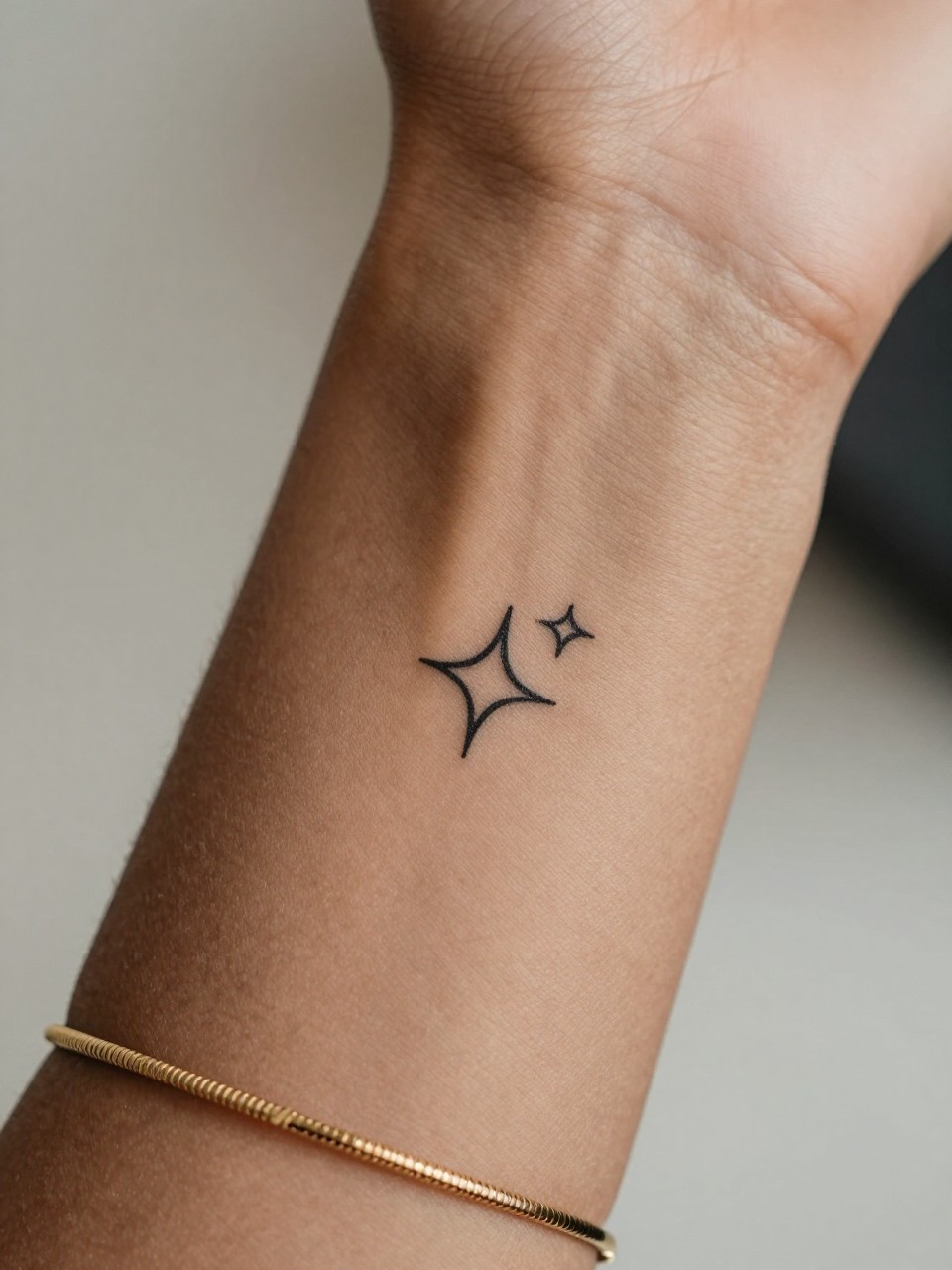 "close-up of a tiny sparkle symbol tattoo on dark skin at the wrist, small black ink design, crisp fine lines, minimalist style, realistic skin texture, delicate gold bracelet, tight crop, soft blurred background, design-focused modern feminine tattoo photography (no watermarks on images)"