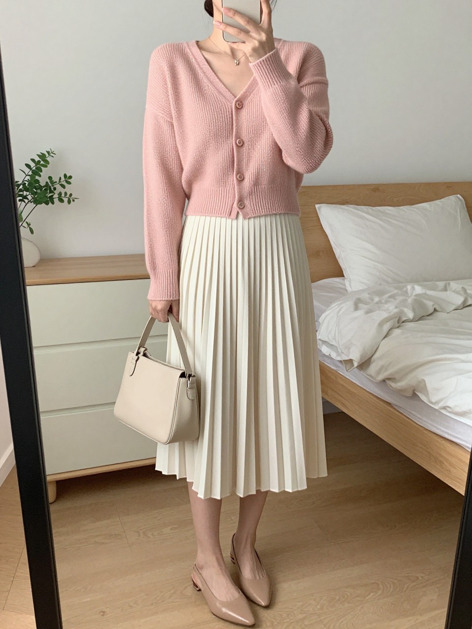 mirror selfie of a woman wearing the exact outfit described above, clearly showing a blush pink knit cropped cardigan, an ivory pleated midi skirt, nude slingback flats, a pale beige leather mini bag, and hair styled in a low bun, holding a phone in front of a mirror, cropped at NECK so the face does not show, with a relaxed casual selfie pose, in a feminine-themed bedroom, soft indoor lighting, including soft pillows on an unmade bed and small plant on a dresser, latest iPhone photo quality. (no watermarks on images)