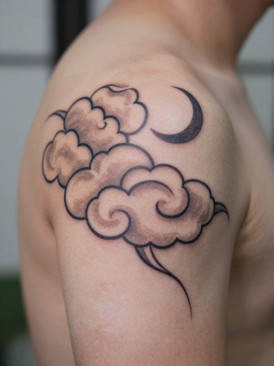 close-up of a Japanese cloud and moon tattoo on the shoulder, traditional cloud forms with a small crescent moon, black and grey ink, clean linework, soft shading, realistic skin texture, design-focused close-up, blurred neutral background, modern tattoo photography (no watermarks on images)