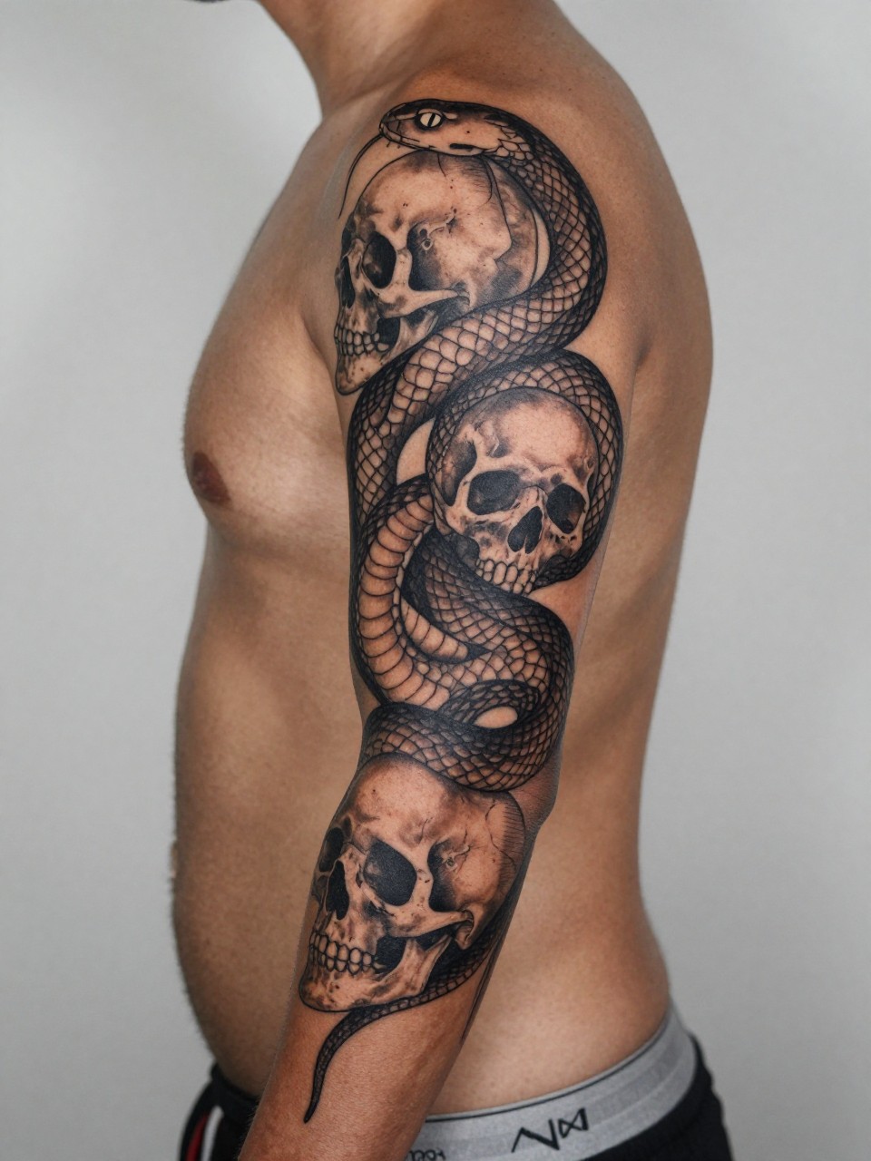 design-focused close-up of a snake and skull full sleeve tattoo on deep brown skin, coiled snake around shaded skulls, black and grey ink, fine scale detail, bold linework, realistic skin texture, tight crop, masculine tattoo styling, neutral blurred background, modern inspiration photo (no watermarks on images)