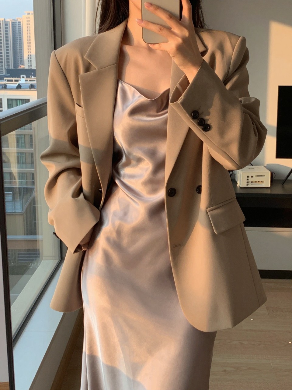 iPhone mirror selfie of a woman in a satin slip dress and oversized blazer, one hand in the blazer pocket, cropped at the neck so face doesn't show, in a city apartment, evening golden hour lighting, iPhone photo quality.