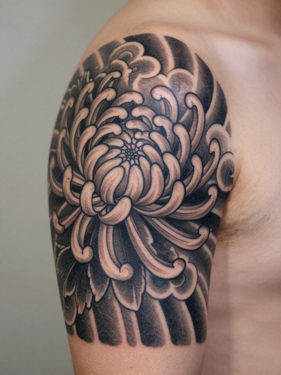 macro shot of a Japanese cloud and chrysanthemum tattoo on the upper arm, layered chrysanthemum bloom with bold cloud detail around it, black and grey ink, crisp traditional linework, soft shading, realistic skin texture, design-first composition, soft neutral background, tattoo inspiration image (no watermarks on images)