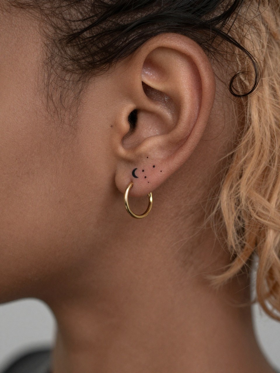 "close-up of a mini crescent moon and dot tattoo on black skin behind the ear, crisp black ink, tiny minimalist design, clean linework, realistic skin texture, small gold hoop earring, tight crop, soft blurred background, design-focused modern tattoo photography (no watermarks on images)"
