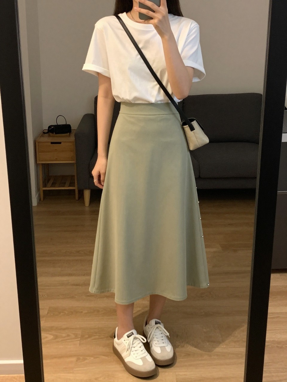 iPhone mirror selfie of a woman in a white tee tucked into a sage green A-line skirt with white sneakers, holding a crossbody bag, cropped at NECK so face doesn't show, in a city apartment, warm moody lighting, iPhone photo quality.