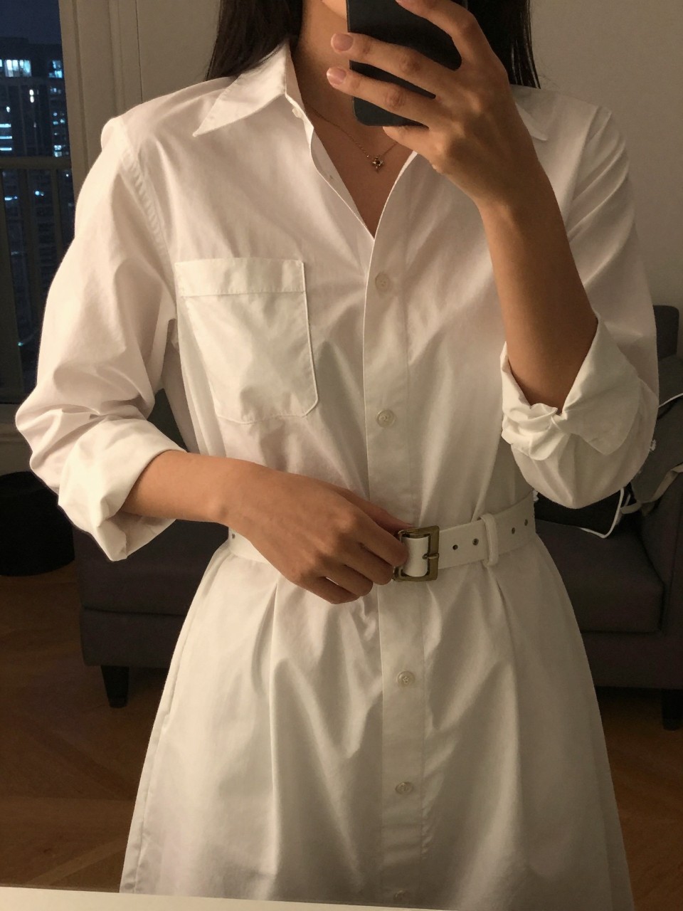 iPhone mirror selfie of a woman in a white shirt dress with a belt, adjusting the buckle, cropped at the neck so face doesn't show, in a city apartment, warm moody lighting, iPhone photo quality.