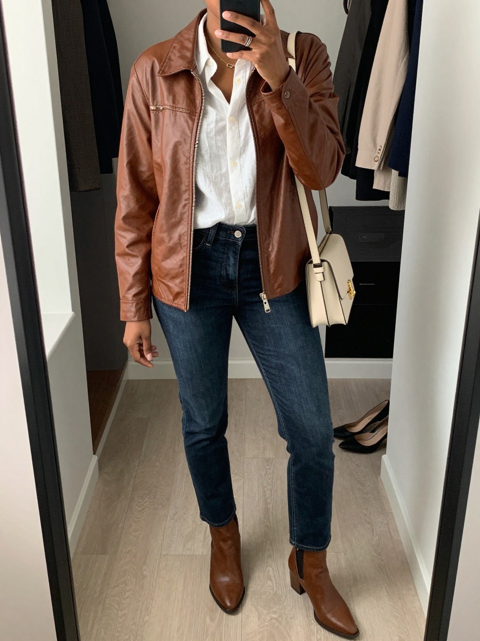 mirror selfie of a black woman wearing the exact outfit described above, clearly showing a soft brown relaxed-fit leather jacket, white linen button-up shirt, dark-wash slim straight jeans, brown ankle boots, and a cream crossbody bag, holding a phone in front of a mirror, cropped at NECK so the face does not show, with a relaxed casual selfie pose, in a walk-in closet mirror, natural window light, including an open closet with clothes visible and a pair of heels on the floor nearby, latest iPhone photo quality.