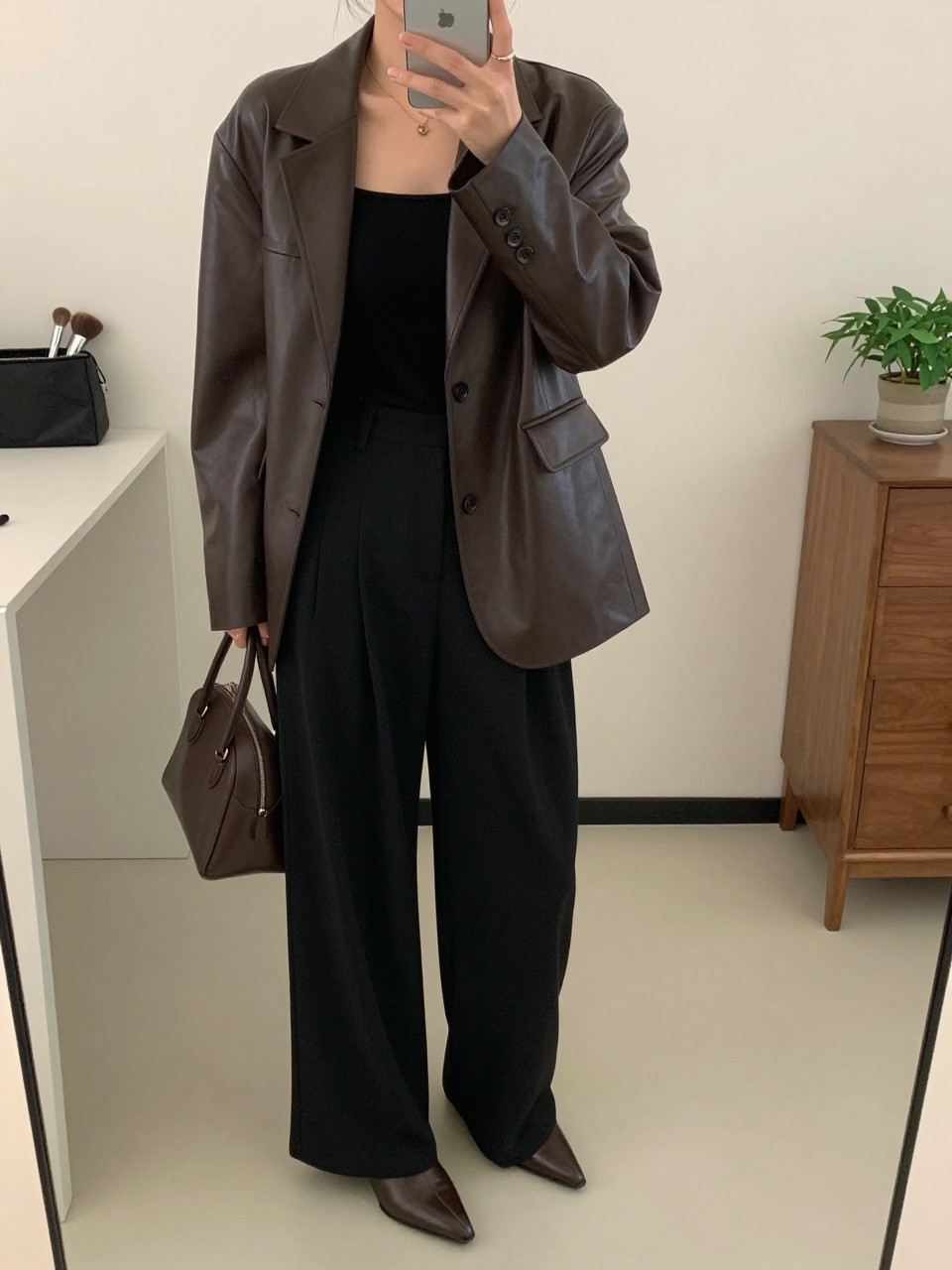 mirror selfie of a woman wearing the exact outfit described above, clearly showing a deep brown polished leather jacket, black fitted square-neck top, black wide-leg trousers with fluid drape, brown pointed ankle boots, and a small dark brown structured leather bag, holding a phone in front of a mirror, cropped at NECK so the face does not show, with a relaxed casual selfie pose, in a minimal dressing room, soft indoor lighting, including a makeup brush or cosmetic bag on the counter and a small plant on a dresser, latest iPhone photo quality.