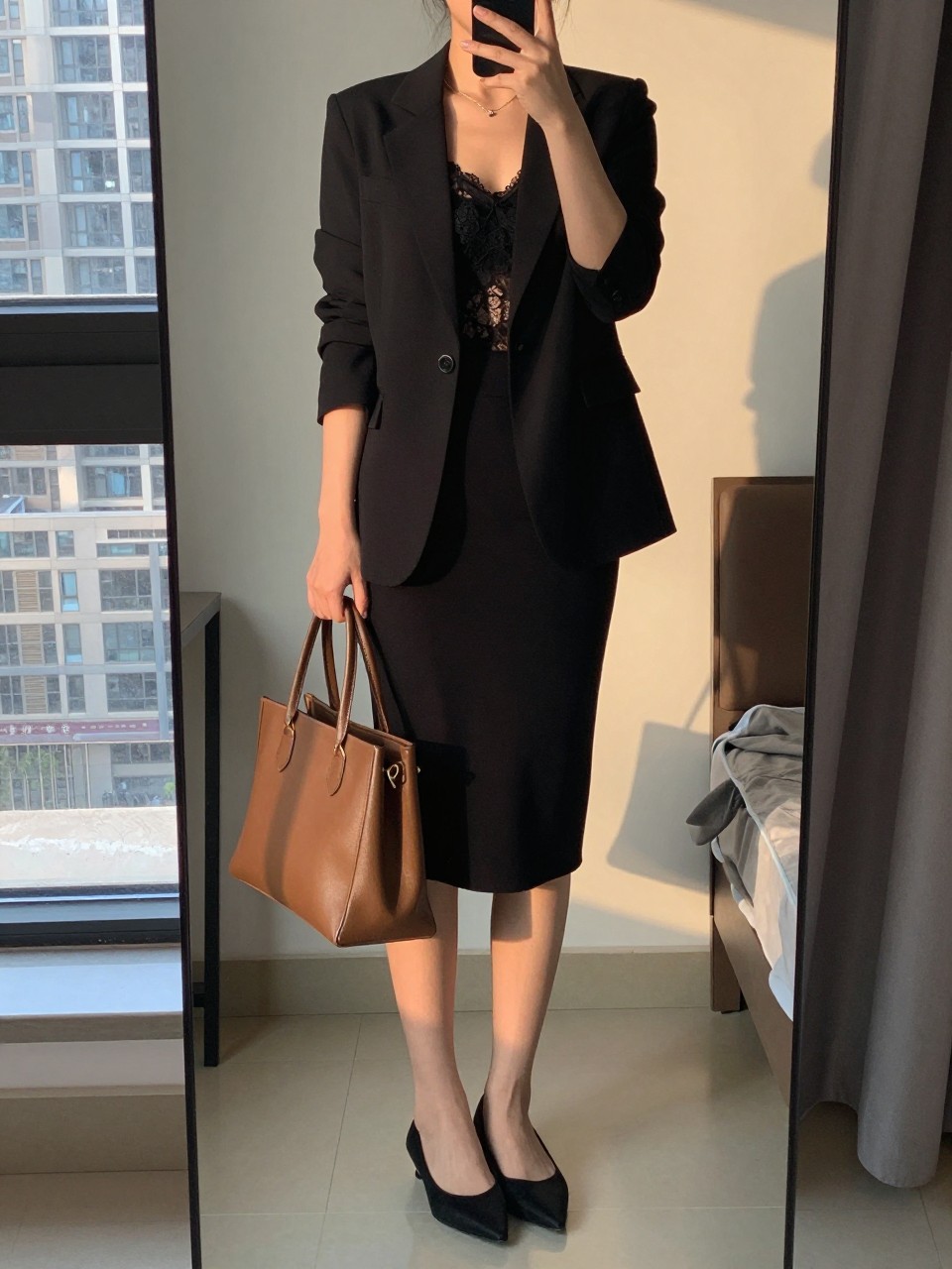 iPhone mirror selfie of a woman in a black blazer over a lace camisole and black pencil skirt with pointed flats, holding a leather tote, cropped at NECK so face doesn't show, in a city apartment, evening golden hour, iPhone photo quality.