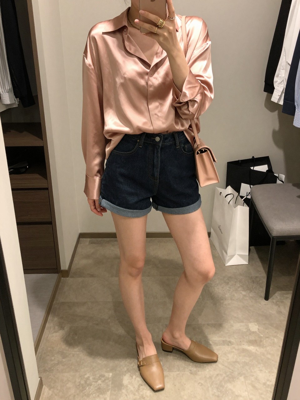 mirror selfie of a woman wearing a rose gold oversized silk shirt with a glossy drape, dark-wash denim shorts with a clean hem, tan heeled mules, a blush crossbody bag, and layered gold rings, holding a phone in front of a mirror, cropped at NECK so the face does not show, with a relaxed casual selfie pose, in a walk-in closet mirror, soft indoor lighting, including an open closet with clothes visible and shopping bags beside a chair, latest iPhone photo quality. (no watermarks on images)