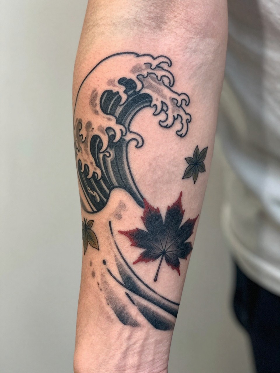 close-up of a clean Japanese wave and maple leaf tattoo on the forearm, curling wave shape with small leaf accents, black and grey ink, crisp traditional linework, soft shading, realistic skin texture, design-focused close-up, blurred neutral background, modern tattoo photography (no watermarks on images)