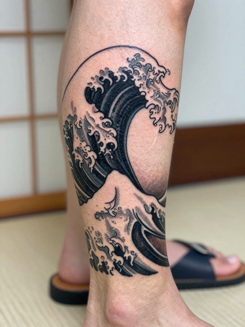 design-focused close-up of a clean Japanese wave tattoo on the calf, bold curling wave, black and grey ink, traditional linework, soft shading, realistic skin texture, sandal strap nearby, minimal blurred background, modern tattoo inspiration photo (no watermarks on images)