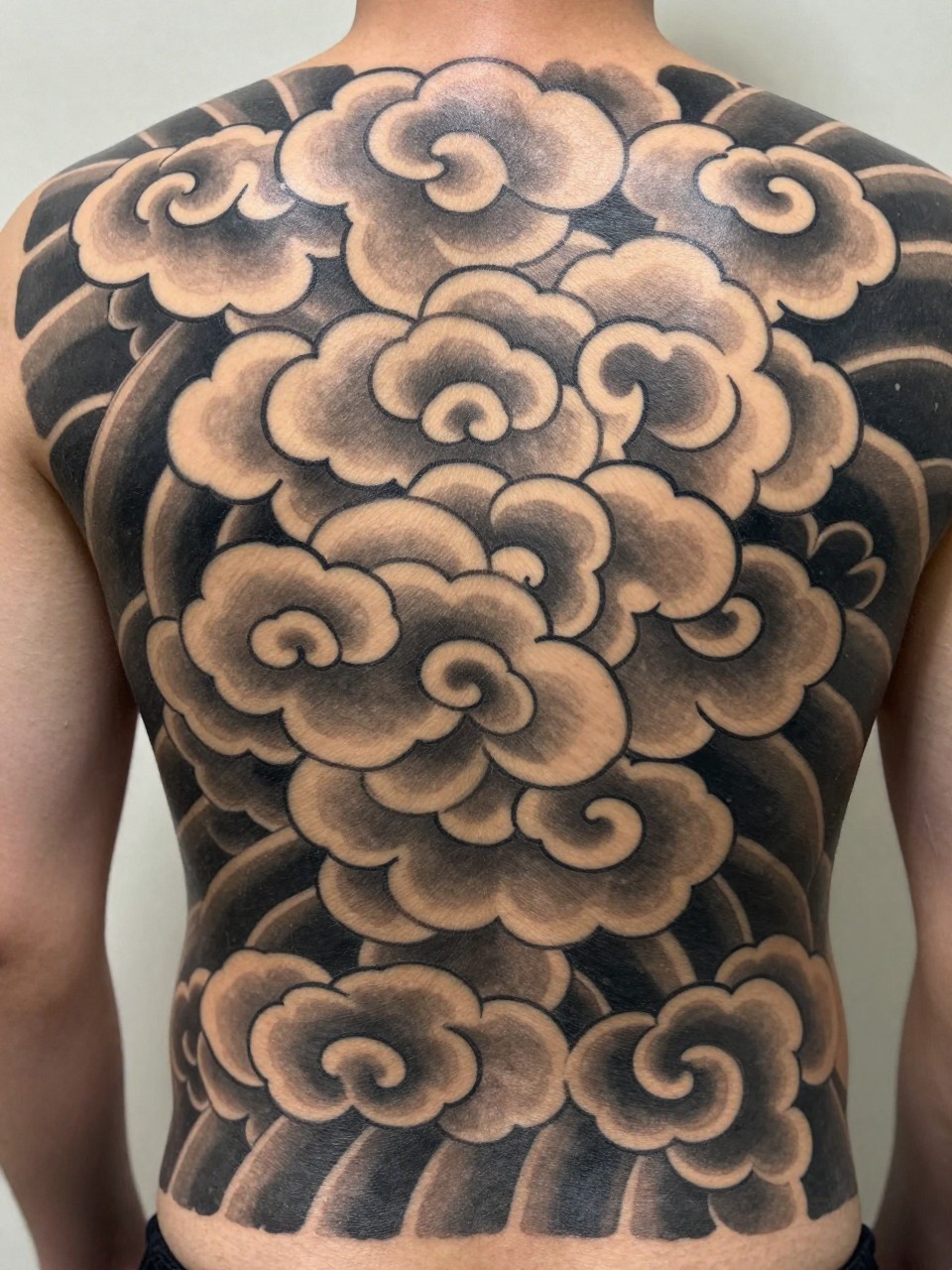 close-up of a full back Japanese cloud tattoo panel, large layered traditional cloud forms across the entire back, black and grey ink, bold linework, smooth shading, strong balanced composition, realistic skin texture, design-focused close-up, soft neutral background, modern tattoo photography (no watermarks on images)