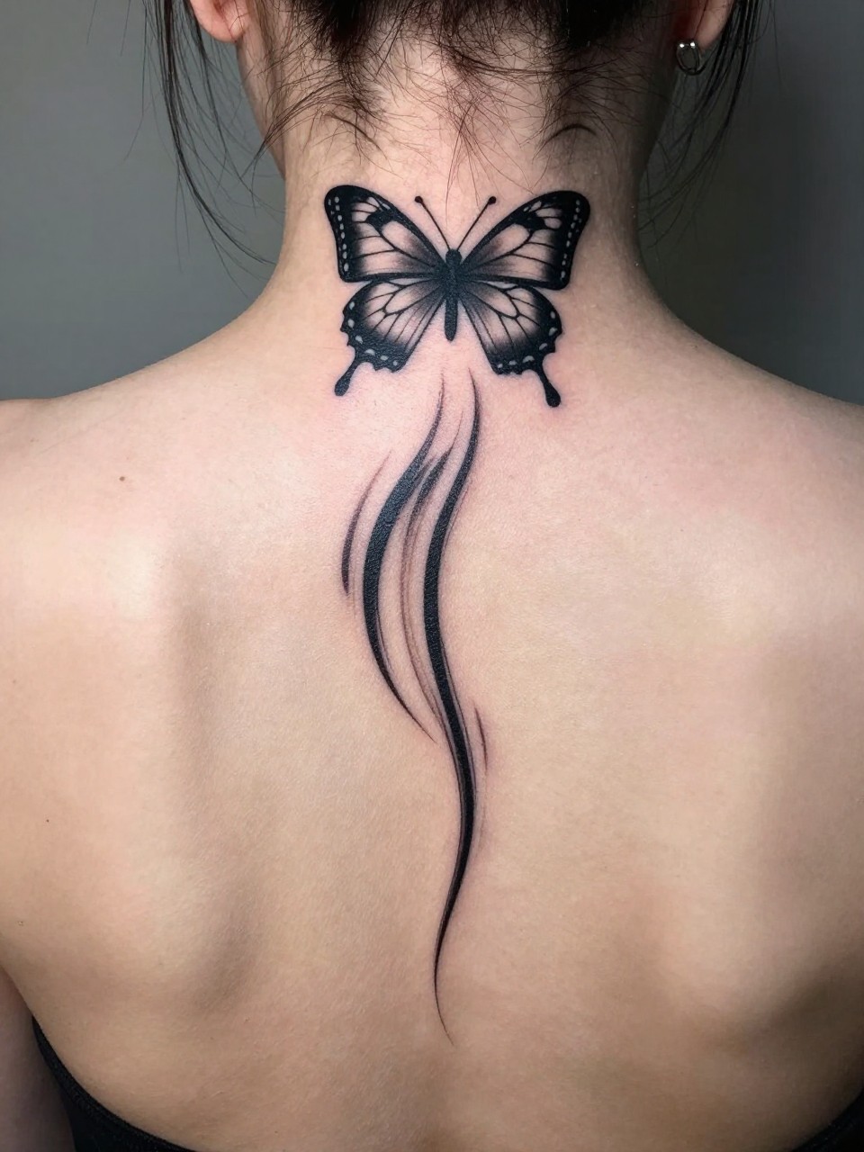"Shot of the upper back and neck: a butterfly tattoo at the cervical spine with bold, sweeping lines trailing down between the shoulder blades. Creates a fierce sense of speed and descent."
