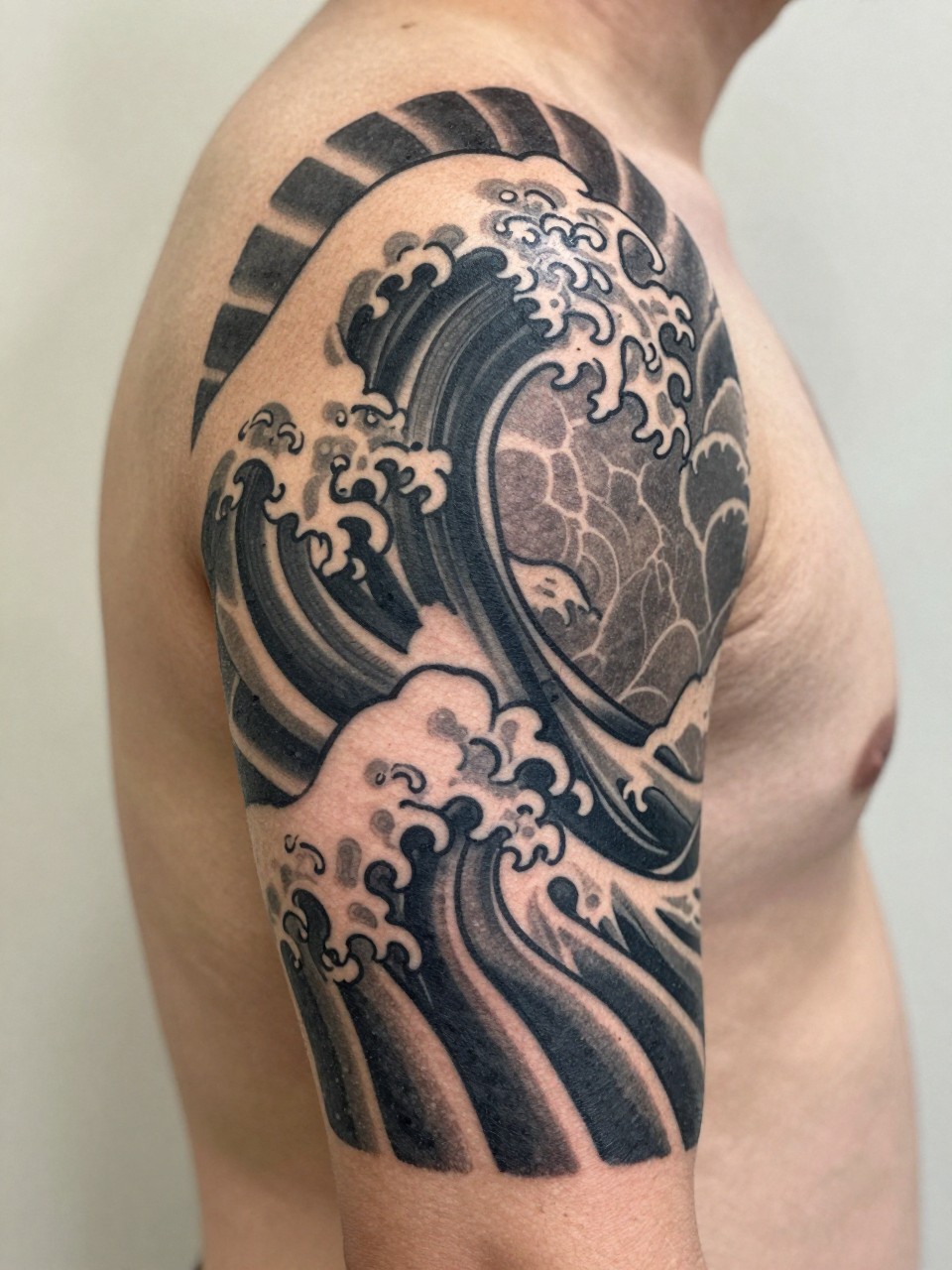 extreme close-up of a traditional Japanese wave armband tattoo on a man's upper arm, repeating cresting wave pattern wrapping around the arm, black and grey ink, bold traditional linework, soft shading, clean masculine composition, realistic skin texture, design-focused close-up, soft neutral background, modern tattoo inspiration photography (no watermarks on images)