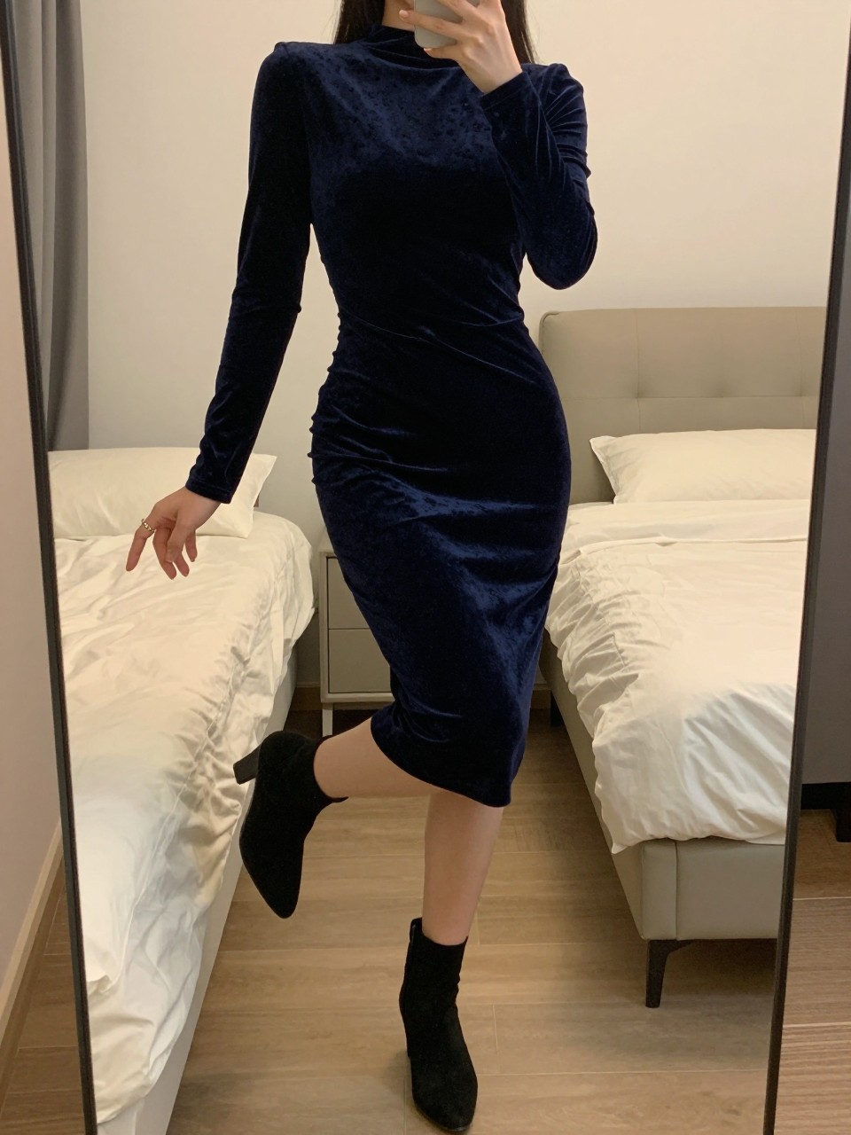 iPhone mirror selfie of a woman wearing a navy velvet bodycon dress with long sleeves and black suede ankle boots, posed with her weight on one leg and arms relaxed, cropped at NECK so face doesn't show, in a feminine-themed bedroom, warm moody lighting, iPhone photo quality.