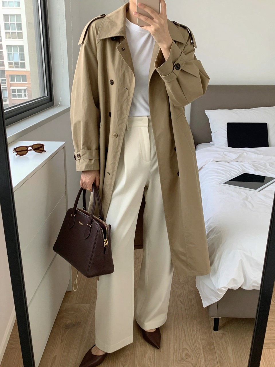 mirror selfie of a woman wearing the exact outfit described above, clearly showing a tan straight-cut barn jacket, white fitted mock-neck top, cream wide-leg trousers in smooth woven fabric, brown pointed flats, and a structured dark brown handbag, holding a phone in front of a mirror, cropped at NECK so the face does not show, with a relaxed casual selfie pose, in a stylish city apartment, natural window light, including sunglasses placed on a dresser and laptop or tablet on the bed, latest iPhone photo quality.
